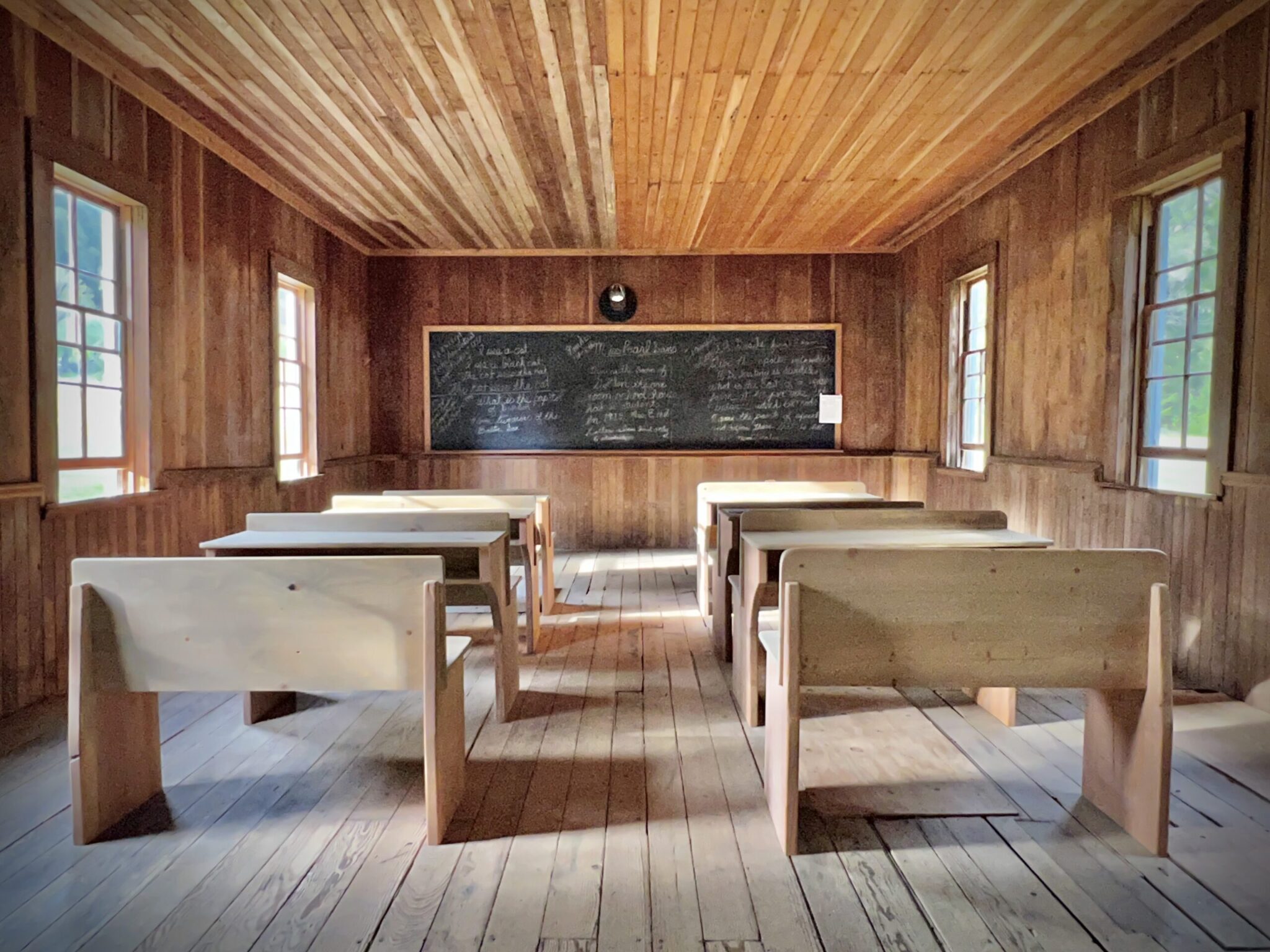 View from the back of the classroom, looking towards the chalkboard, everything in the room besides the chalkboard and glass windows is entirely wooden. There are two rows of seats leading up to the board, they are benches with desks built into the back of them, and there is illegible writing all over the chalkboard.