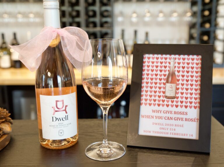 A bottle of wine from Dwell and a half-filled glass are on a bar counter beside a sign reading, "Why give roses when you can give rose'?" In the background, more wine glasses hang upside down against the wall, and additional bottles are on the counter.