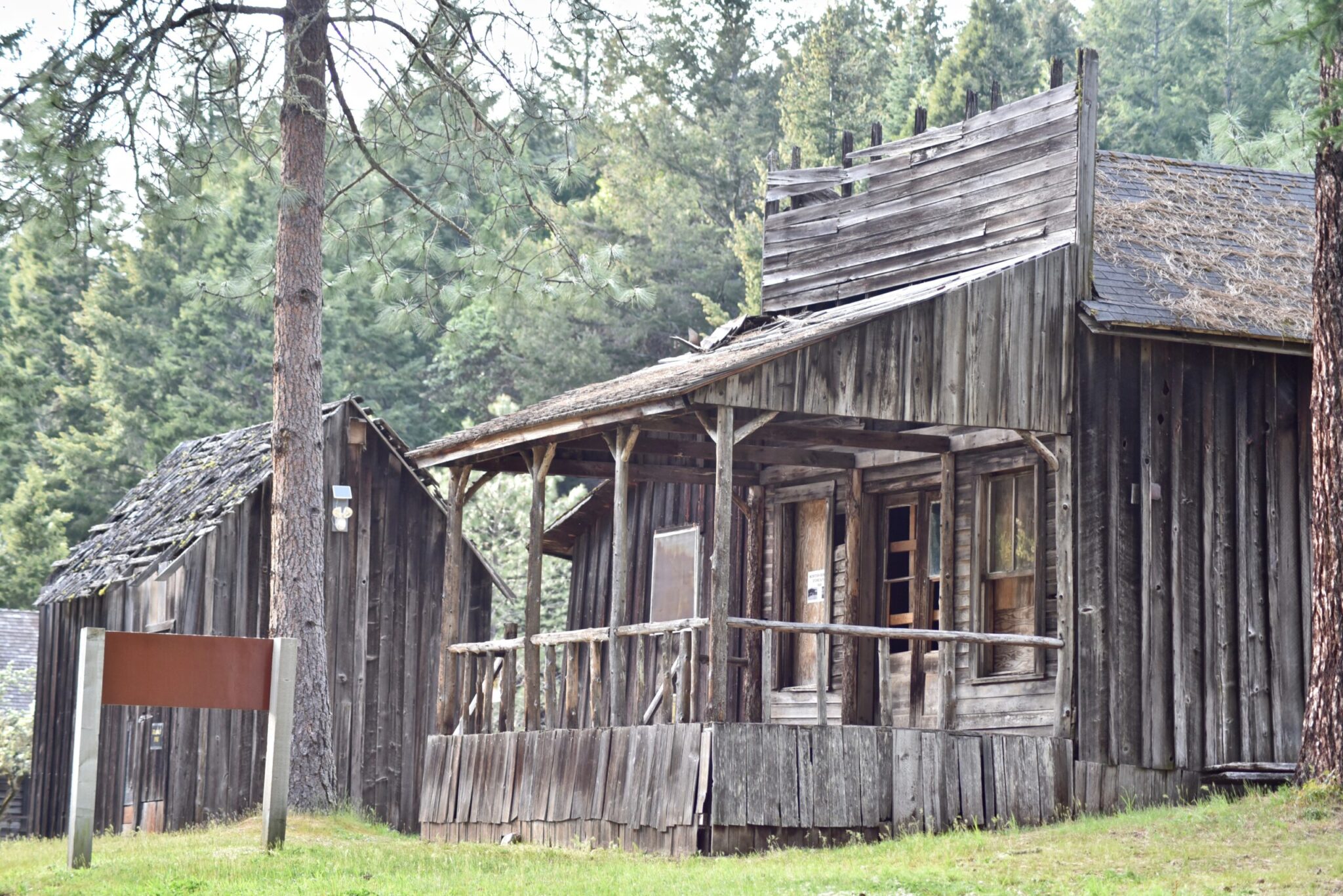 Side glimpse at the ghost town in Southern Oregon, focusing on a small abandoned store with the old square sign in front of the roof and the porch protruding out the front. To the left of the building, there is a tree, and behind it, a smaller structure appears to be a tiny wooden outhouse.
