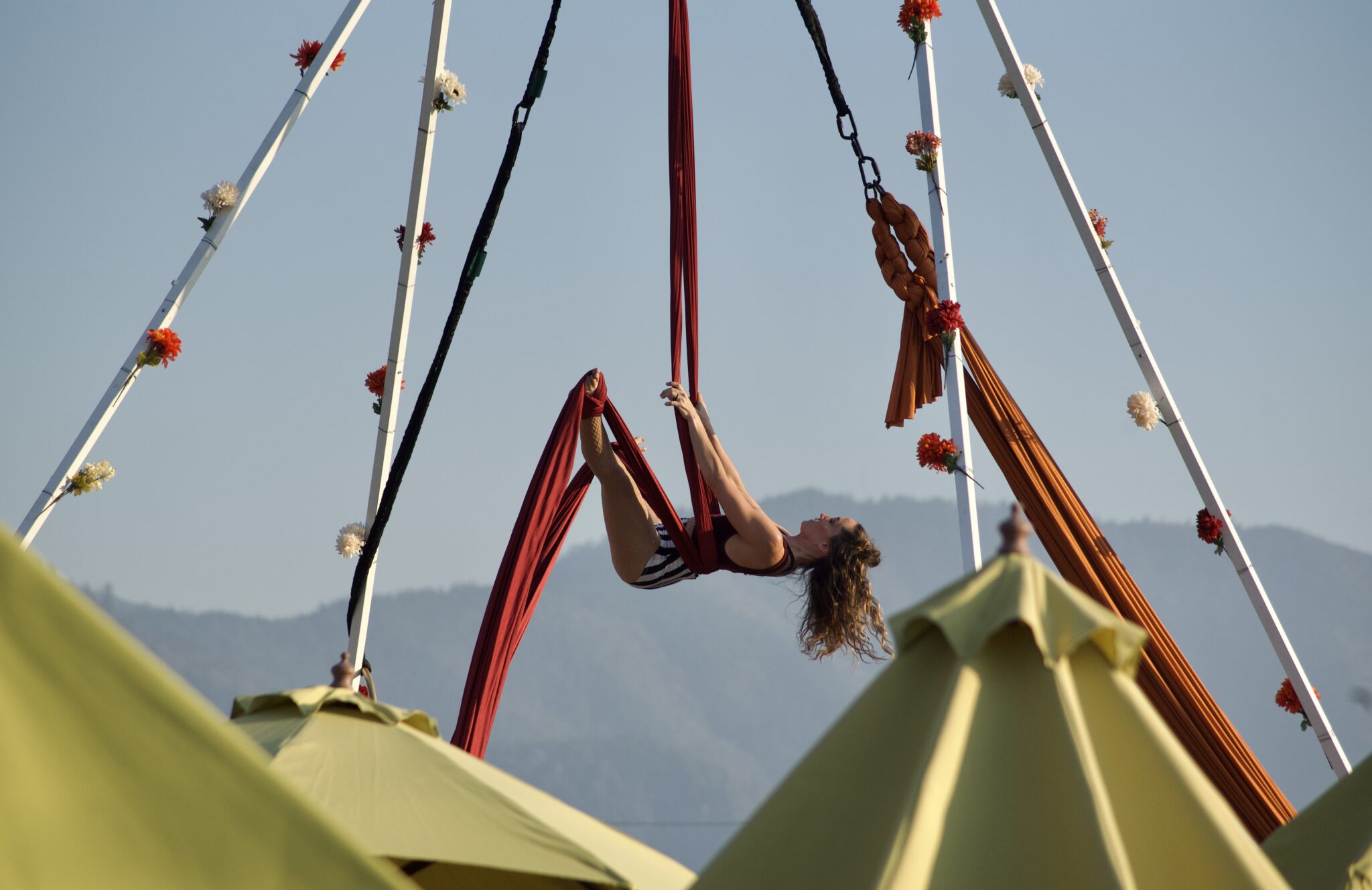 Ethereal Rebels, a gymnast, is suspended in midair outdoors; she is spinning, wearing a black and white striped onesie, handling from two large red ribbons.