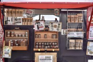 Storefront of a farmers market stands with locally made products in jars and small wax bags. There is a little window in the middle of the stand with a sign that reads "Tea."