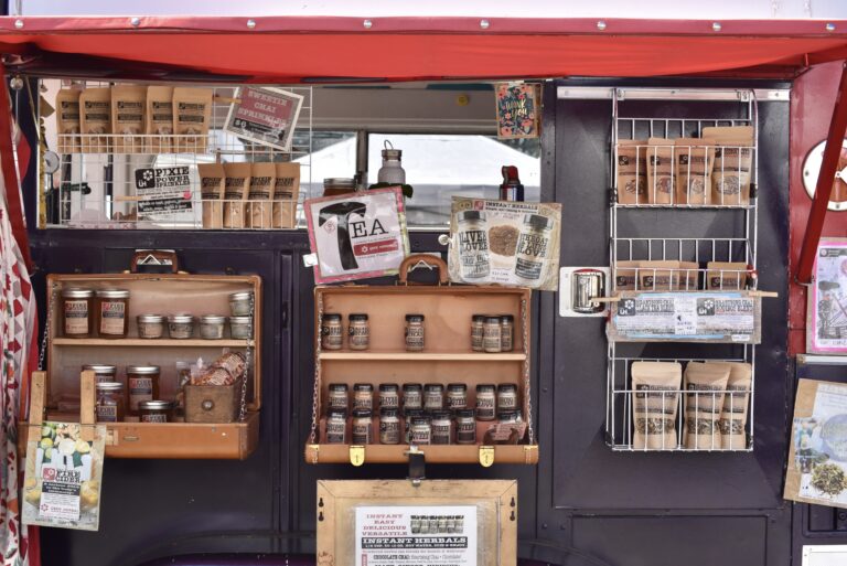 Storefront of a farmers market stands with locally made products in jars and small wax bags. There is a little window in the middle of the stand with a sign that reads "Tea."