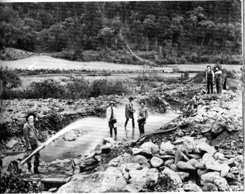 Vintage photo of a group of people standing on a rock formation and some in a puddle of water as another spray a hose at them.