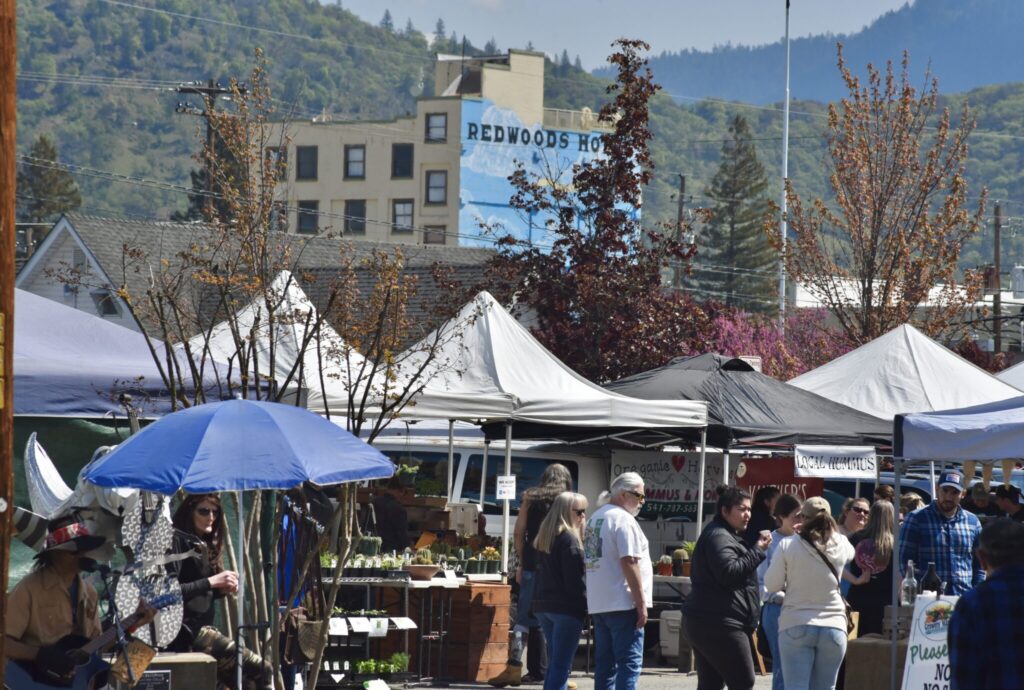 There is a busy street full of market vendors with tents; in the background, there is a glimpse of downtown with the most significant building with a large mural painted on the side. The wall reads "Redwood Hotel."