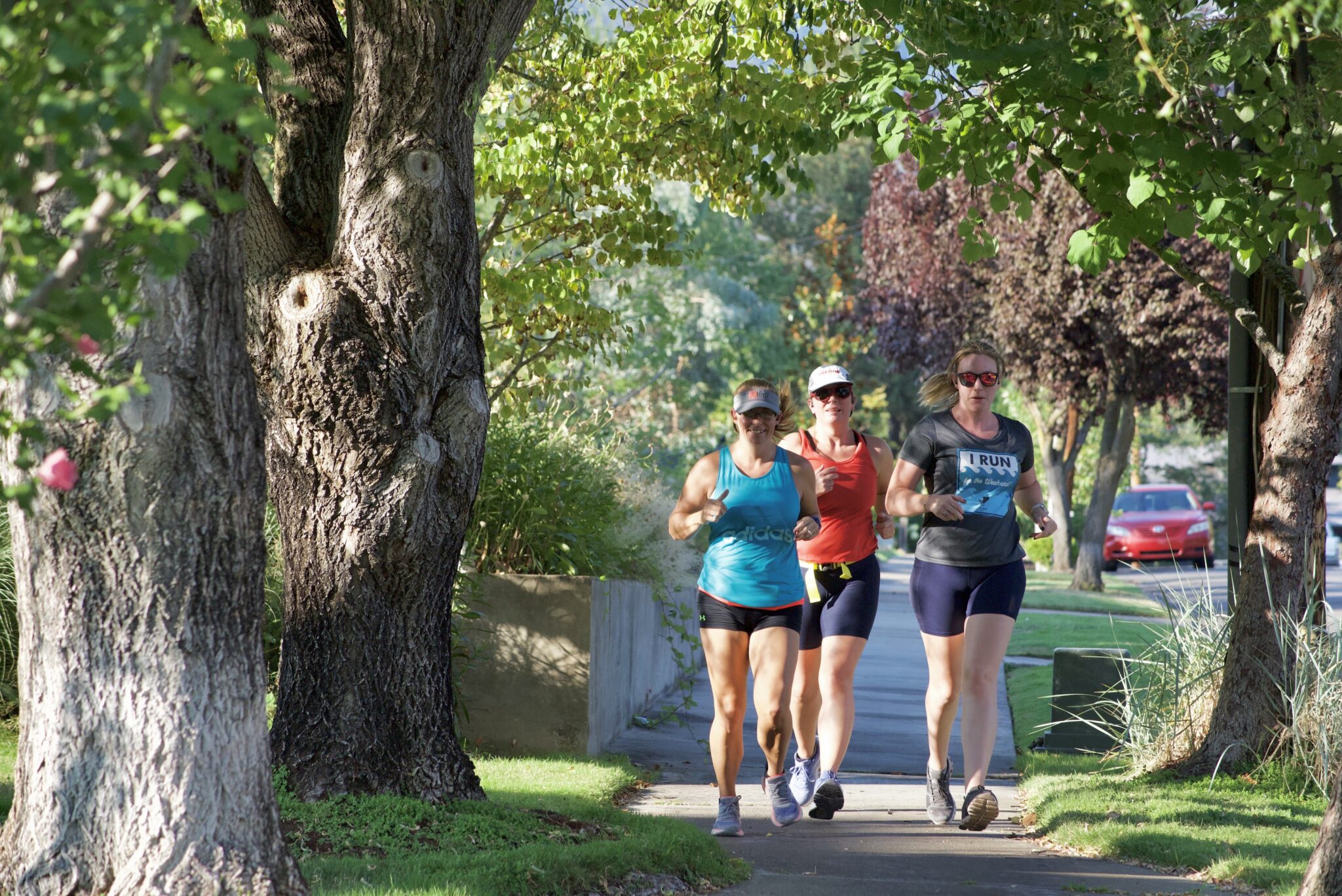 Three women are jogging together down a sidewalk towards the camera; there are trees creating a frame around them on both sides.