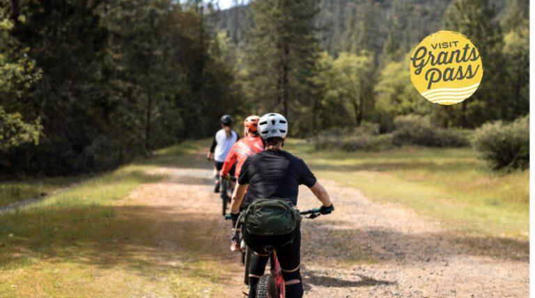Three cyclists are going down an outdoor path away from the camera. The trail is on an open field leading into a cluster of trees; in the top right corner is the Visit Grants Pass logo.