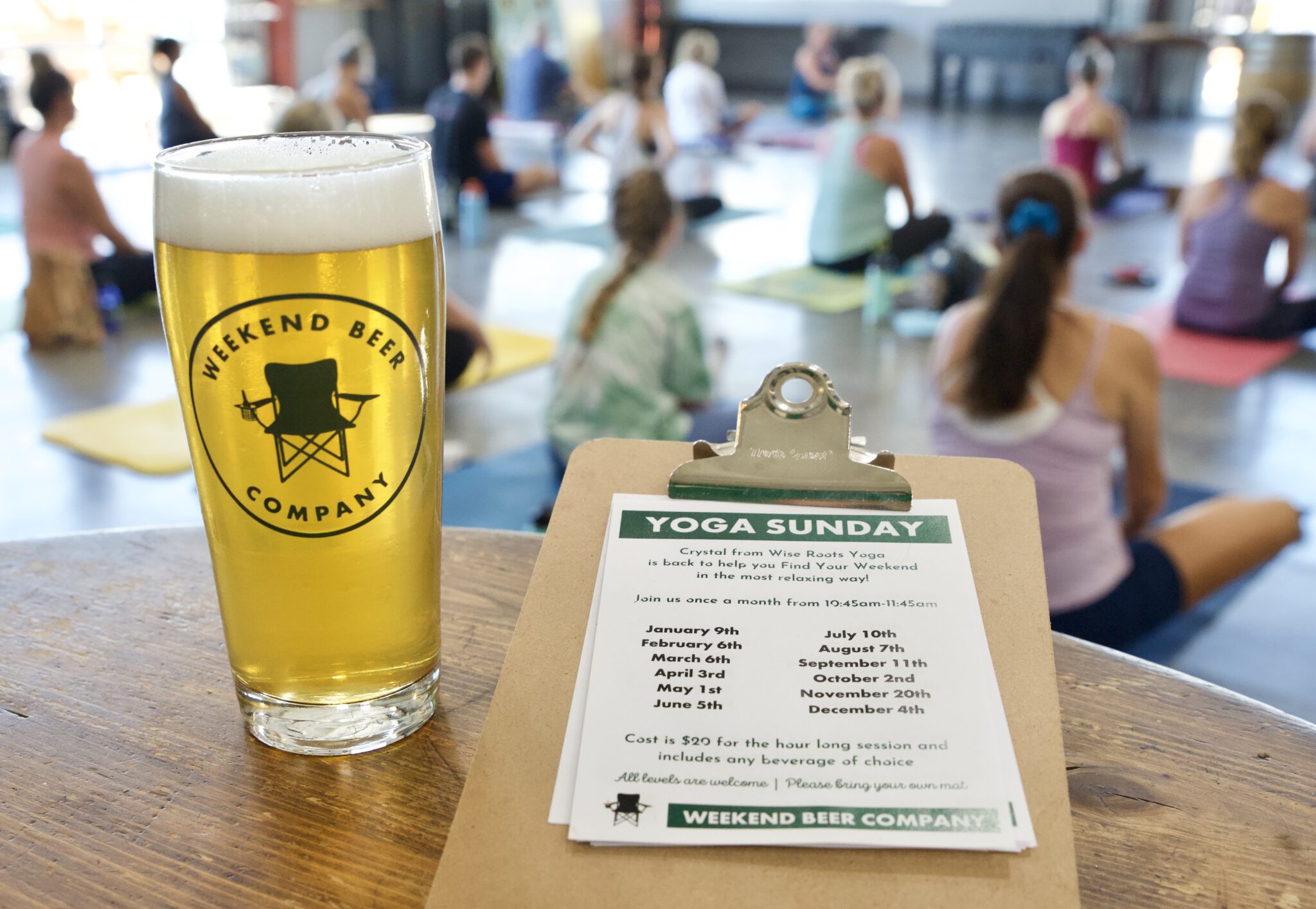 A yoga class can be seen sitting in a well-lit room; they in the background are slightly blurred. In the foreground, there is a table edge, a beer glass with the logo "Weekend Beer Company" sits on the right side, and on the left, there is a clipboard reading "Yoga Sunday" listing dates.