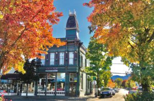 Fall trees create a frame around one of Grants Pass downtown historic buildings, a tall blue Victorian style with a tower-like room and elegant details.