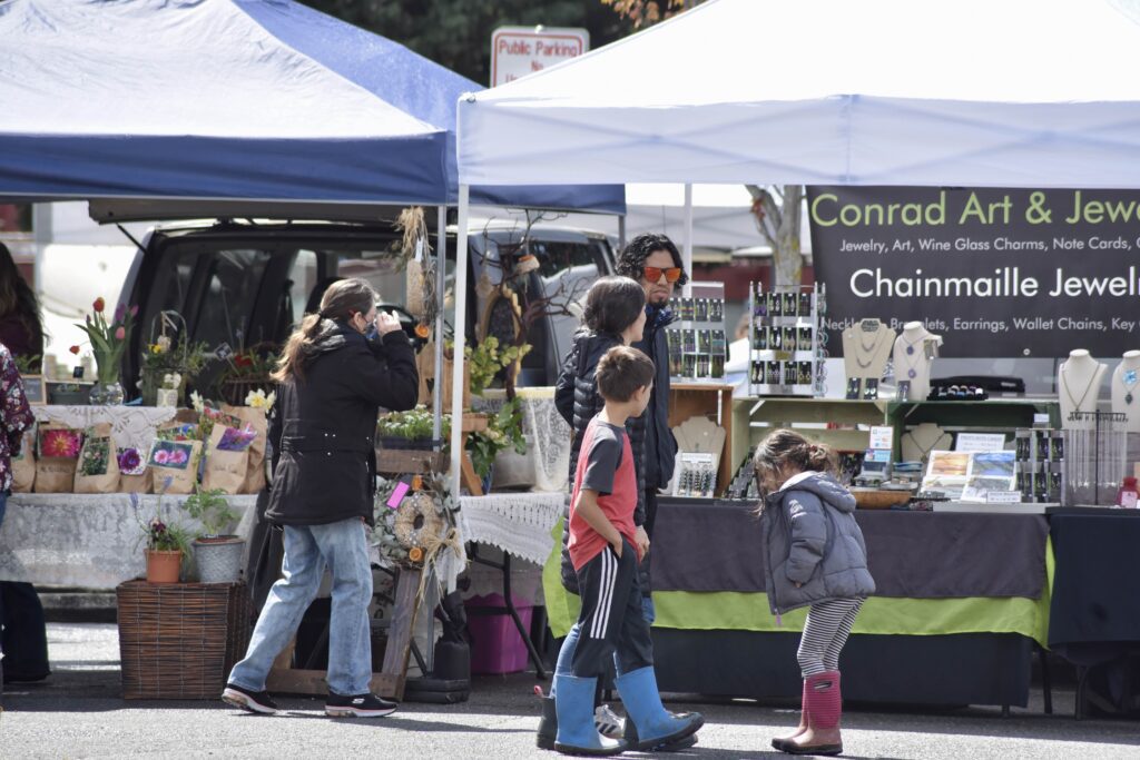 A family walks by the storefont stands set up at the Farm to Fork market. There are boutique items and flowers on display.