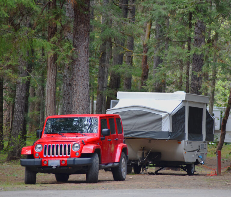 A bright red Jeep Wrangler is backed into an RV camping spot; it has a trailer attached that has been popped up with canopy fixtures sticking out. In the background, tall tree trunks can be seen, with the tree tops not visible.