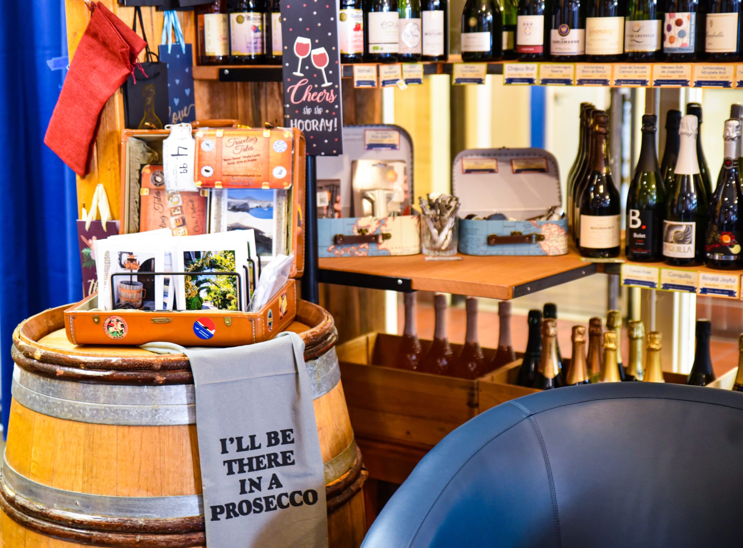 There is a small boutique store in a wine shop featuring memorabilia for wine country, nicely presented on top of a wood barrel, in front of shelves of local wines.