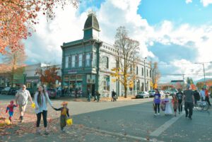 Corner view of one of the historic buildings in downtown Grants Pass. The streets in front of the building are full of pedestrians walking around in costumes with their Halloween candy. The sky is cloudy with a patch of bright blue shining through, and the colors of fall are shown throughout the leaves on the trees and the ones that have failed onto the sidewalk.