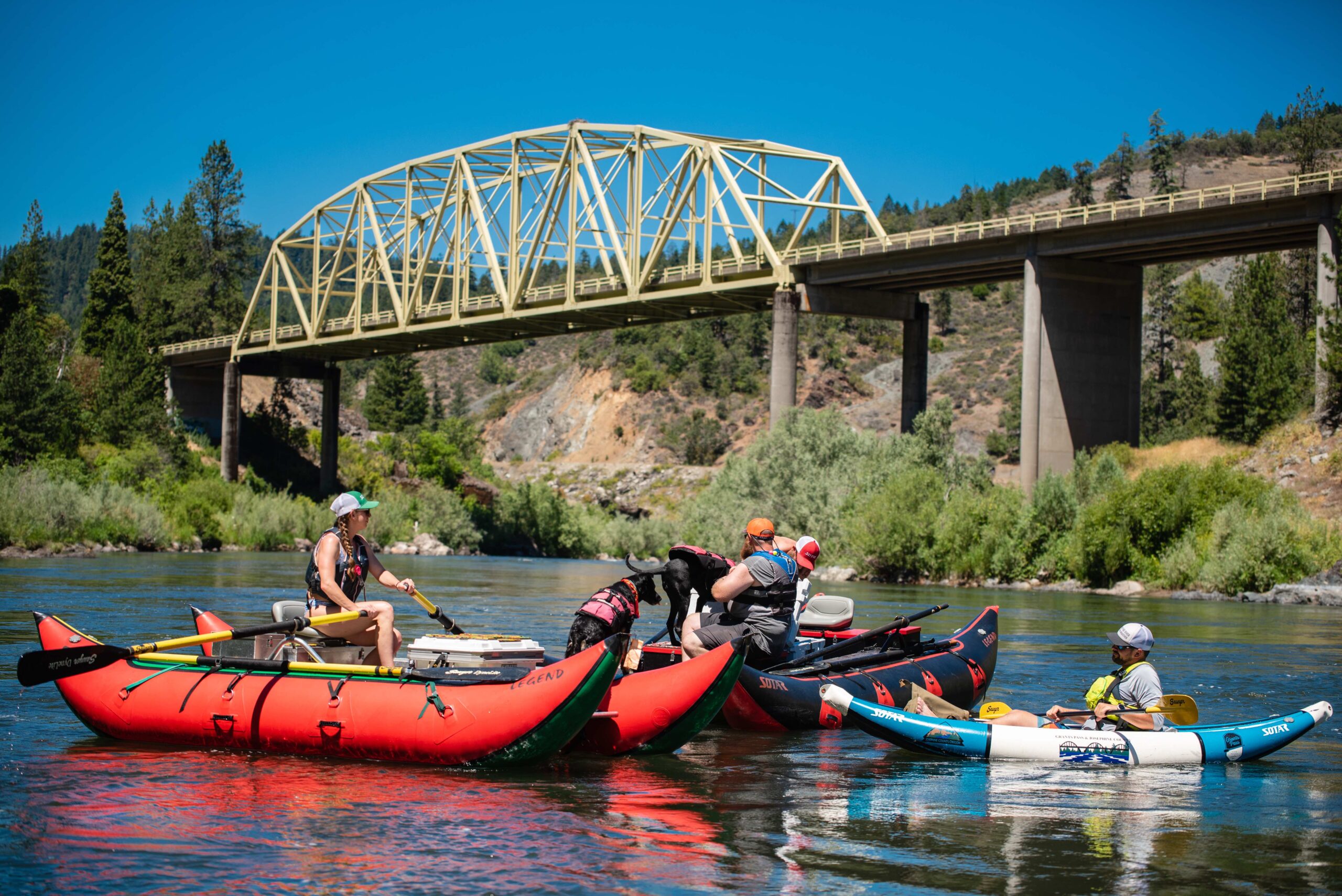 Passing under one of the many bridges in Grants Pass, a person, and their dog drift on a pontoon down the Rogue River. A man in a kayak is also approaching the float on the right.