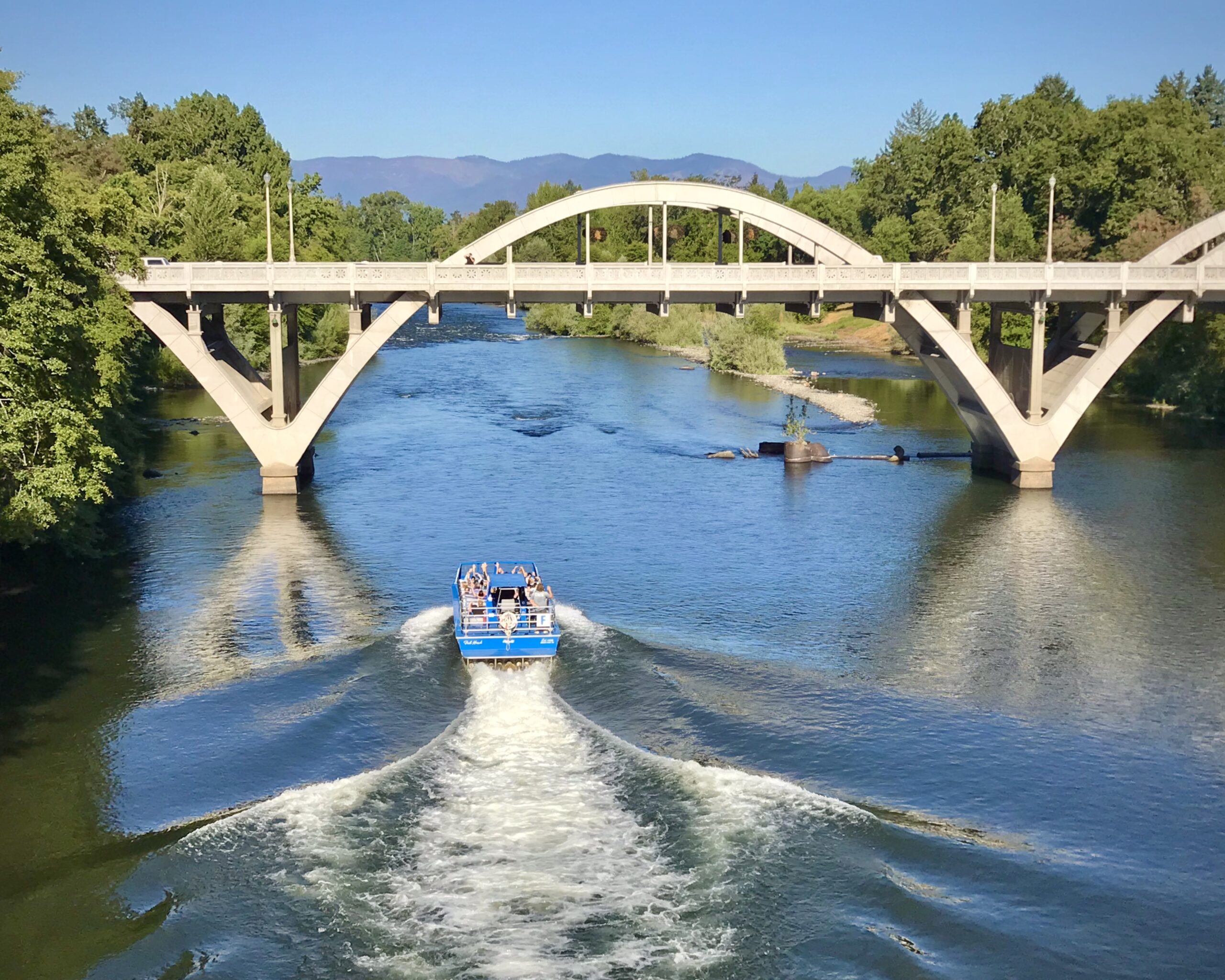 The Hellgate jet boat is tearing through the water, leaving vast ripples behind it as it zooms towards passing under the bridge to Grants Pass, heading down the deep blue Rogue River.