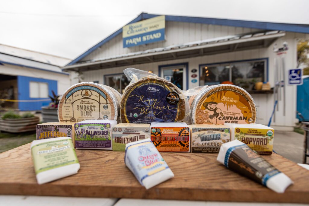 An assortment of local cheeses are on display on. a wood board, showcasing the Smokey Blue, Rogue River Blue, and Caveman Blue. The Creamery can be seen in the background.