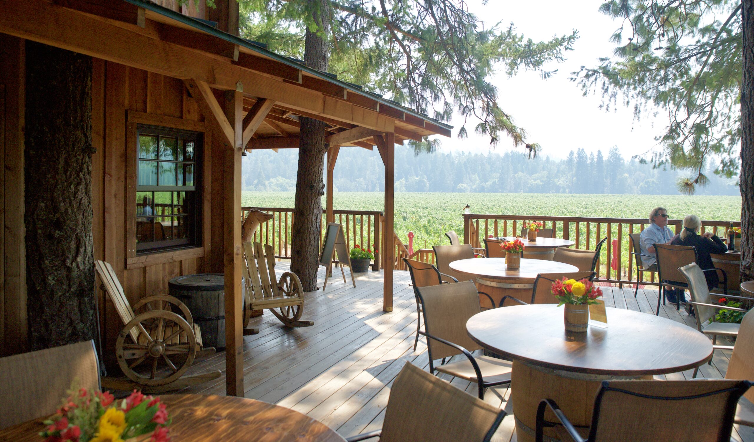 A couple sits on a vineyard patio that wraps around a nice cabin, there are old western chairs, and the patio is built around the trees.