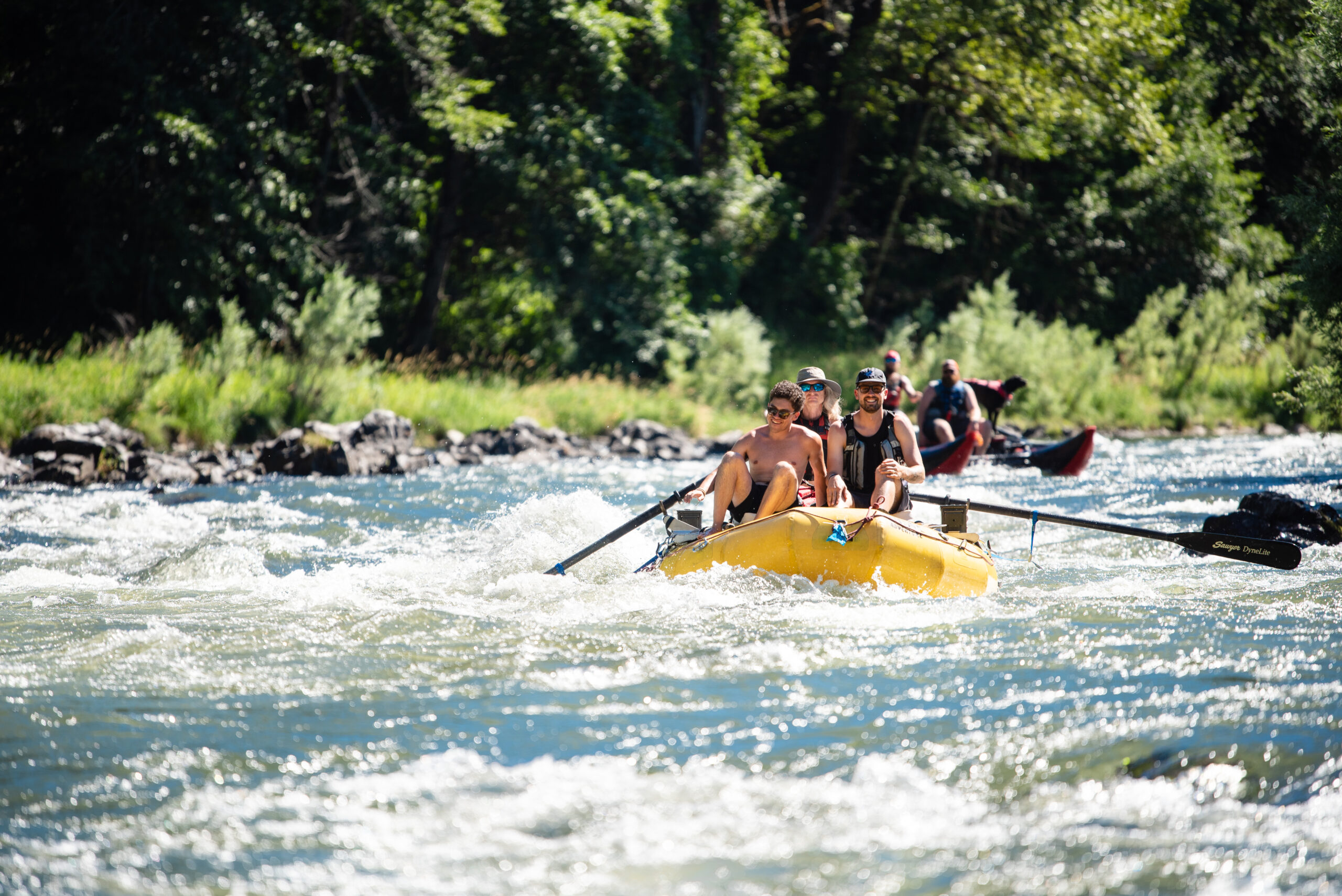 A raft is approaching rapid waters on the Rogue River, it is a small yellow raft with two paddles and three riders. In the background there is a pontoon with two people and their dog in a lifejacket.