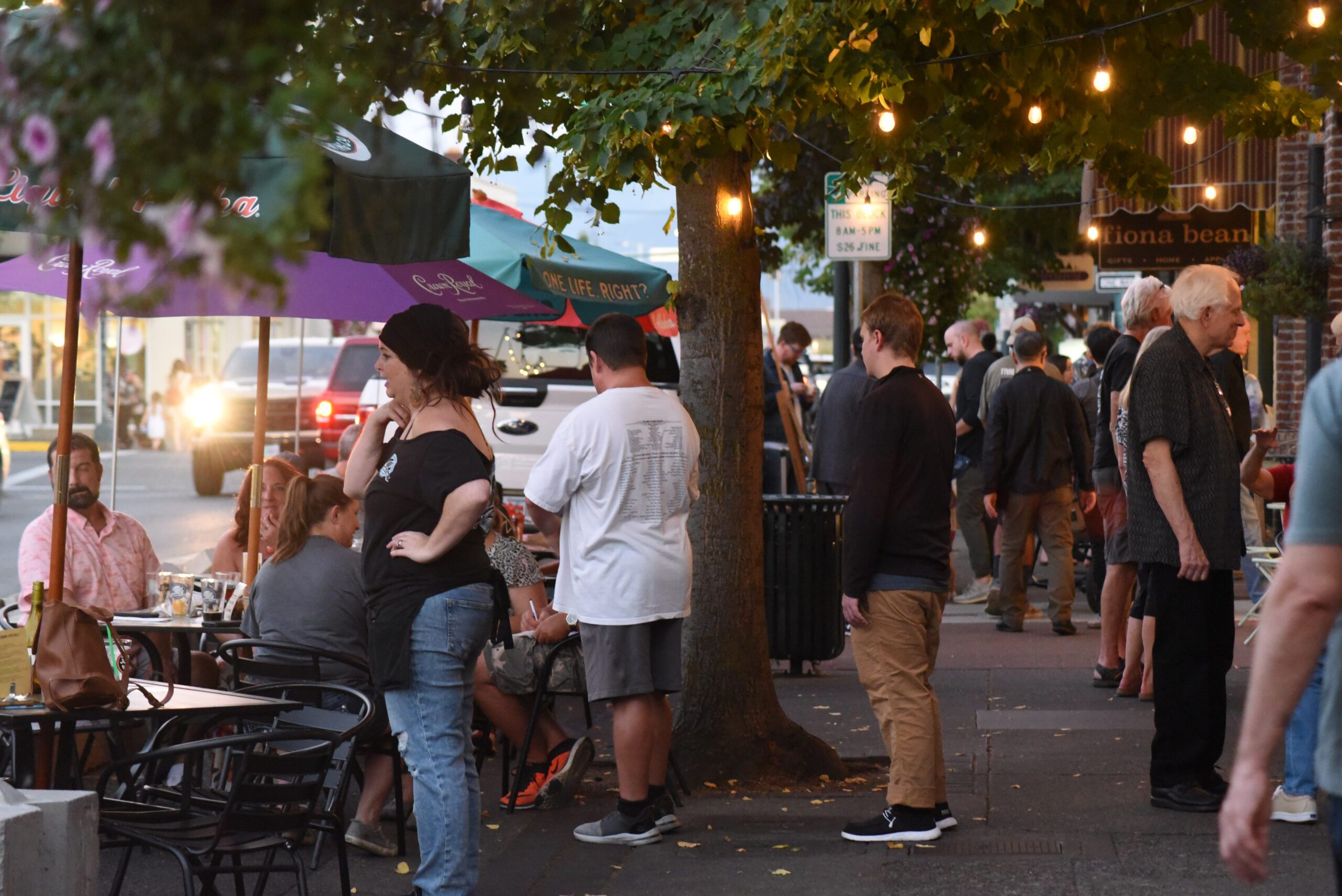 There is a busy street market, it appears to be dusk, but still light out, complimented by the glow of string lights. There is a crowded sidewalk with pedestrians and vendors in downtown Grants Pass.