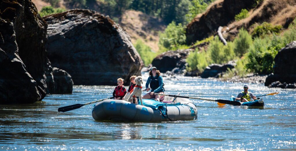 A raft with a parent, two kids, and a dog, floats down the Rogue River, as a kayaker trails behind.