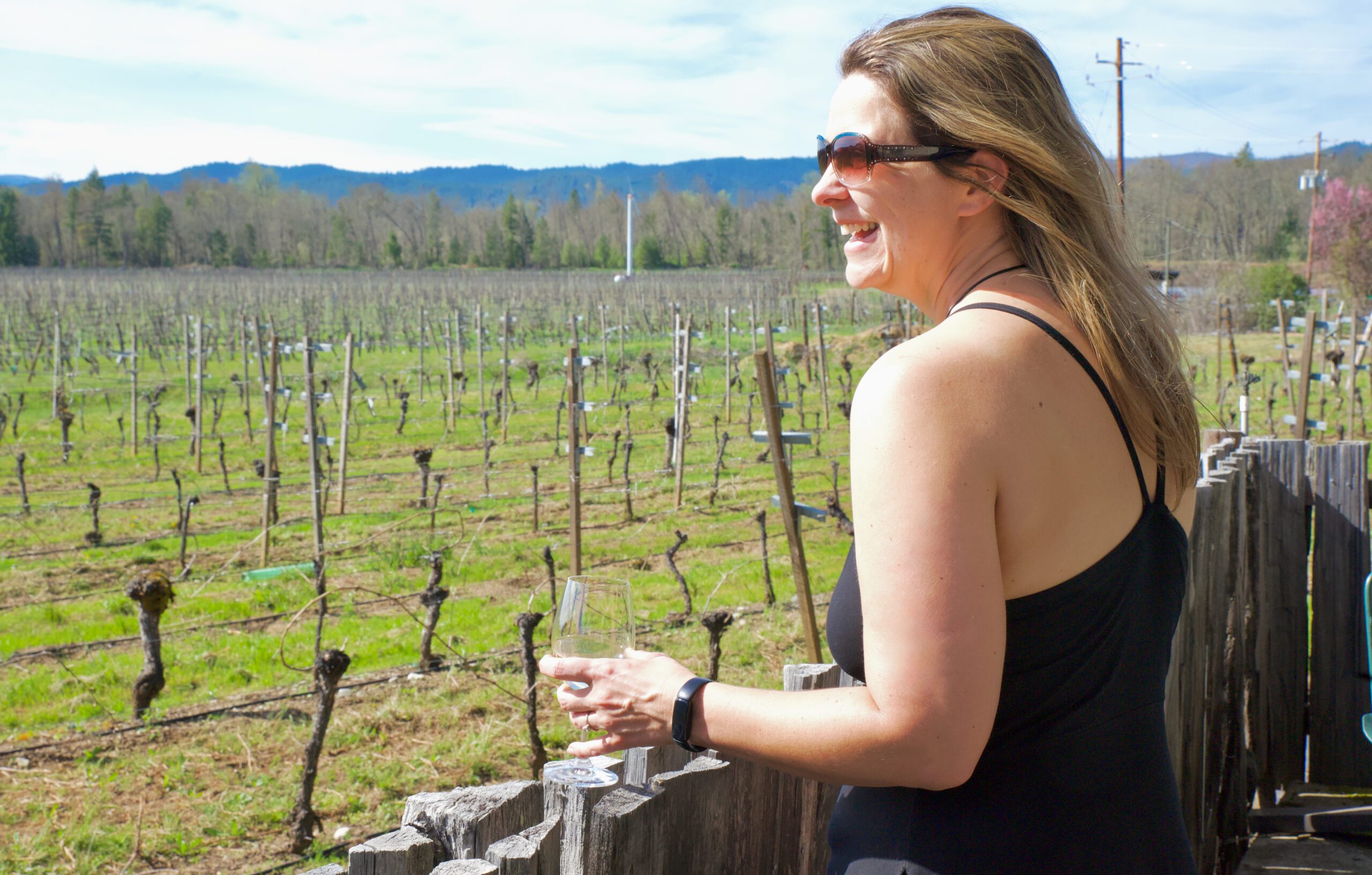 There is a person looking out across a vineyard, laughing while holding a cup of wine, enjoying the view.