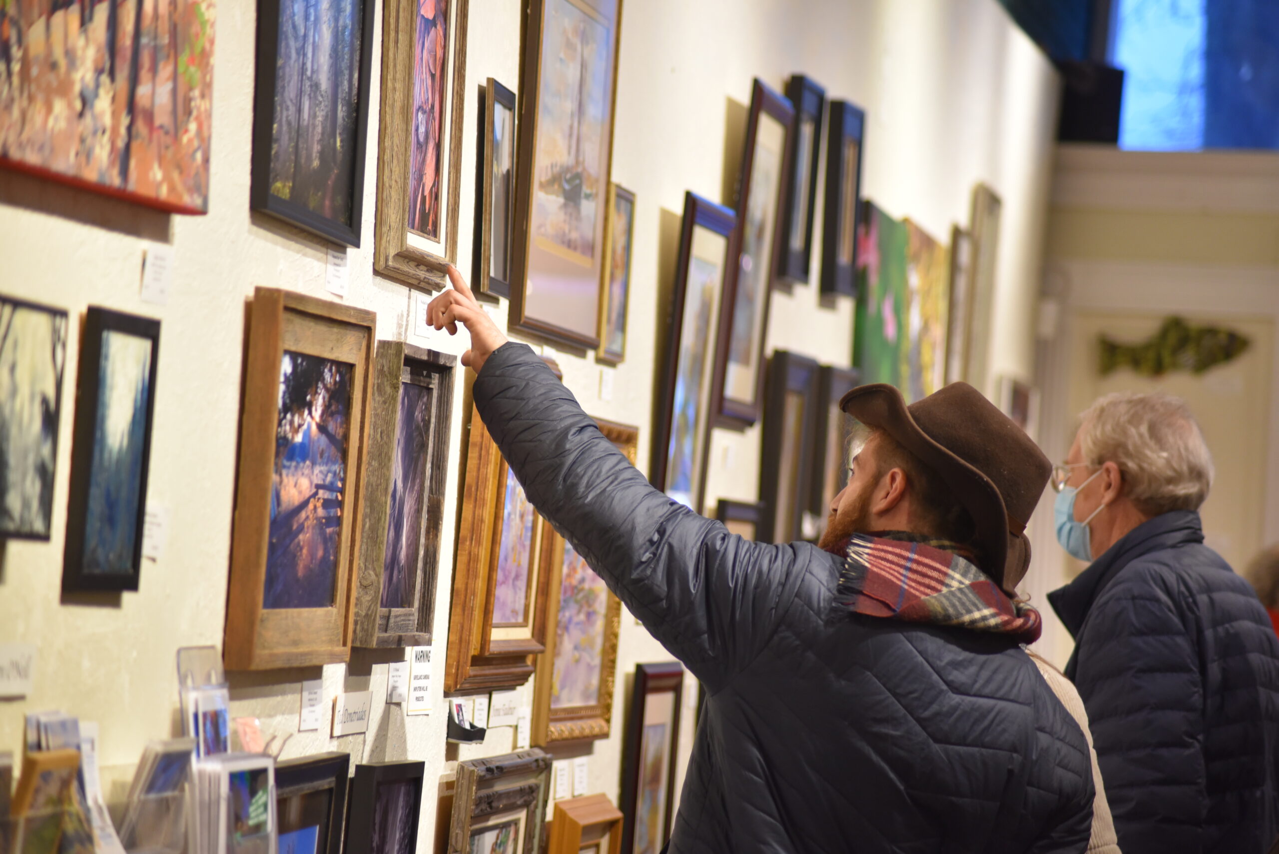 Gallery with viewers, the wall is covered in framed pictures of a variety of styles of art, at the Grants Pass Museum of Art.