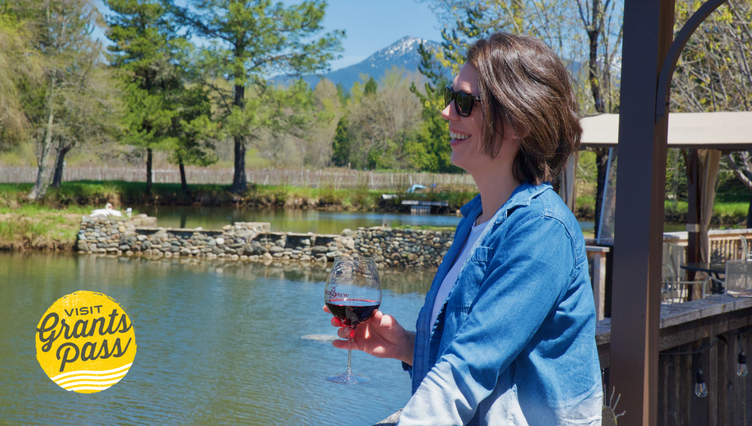 Cover of the Oregon Wine Month featured blog, a person laughs as they hold a glass of wine standing by a creek, there is a tree line in the background with a mountain peeking over.