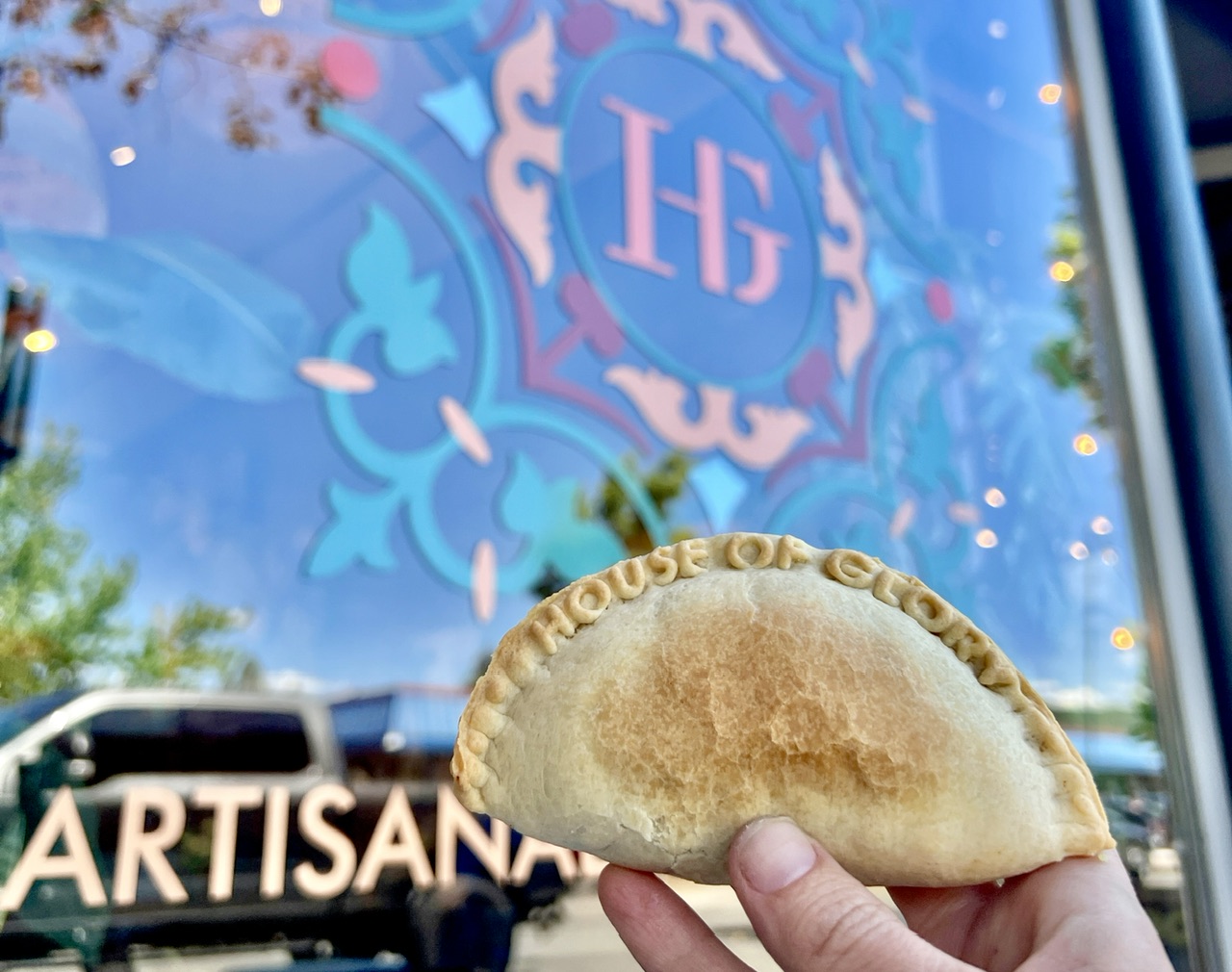 House of Glory dumpling being held up in front of the window sign, with the letters HG surrounded by elegant shapes and designs. The dumpling has the words House of Glory imprinted on it.