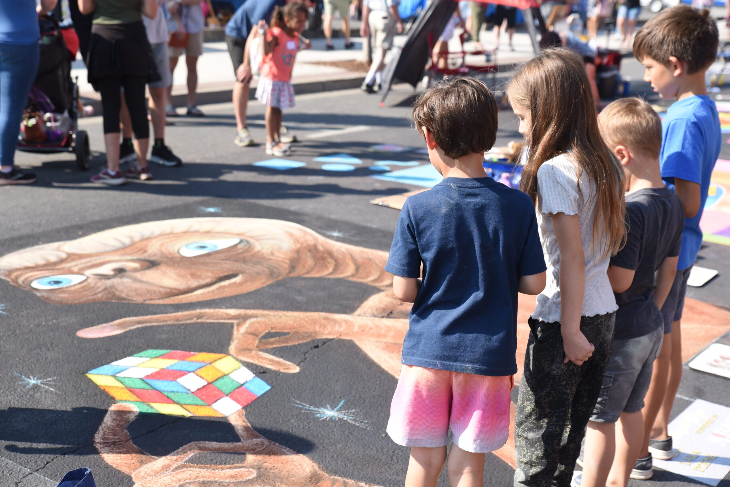 Four kids are lined up admiring the chalk art of a design of ET the alien with a rubik's cube.