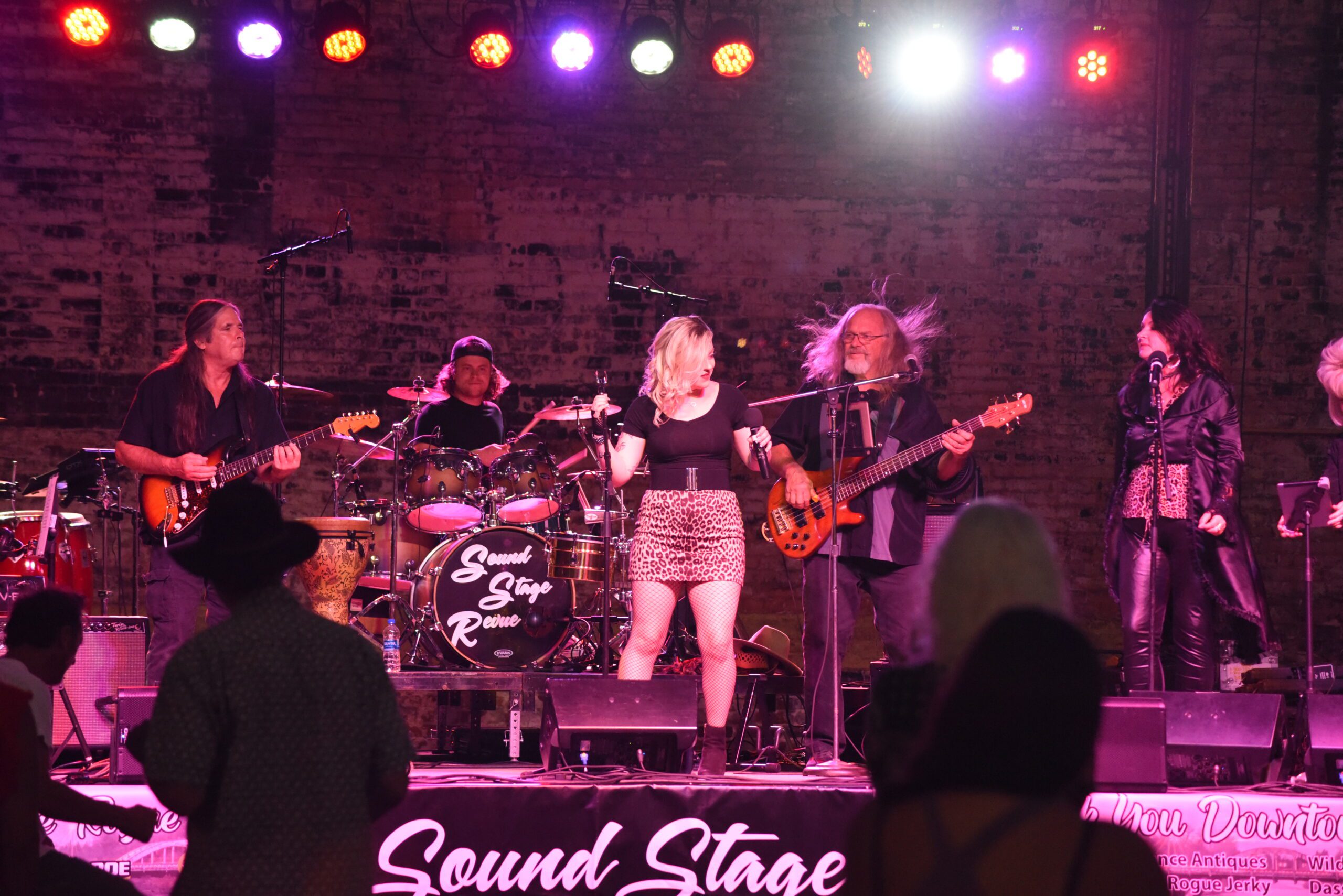 Five band members are playing live music, two singers, two guitarists, and a drummer. They are in front of a brick wall and under glowing purple and red lights.
