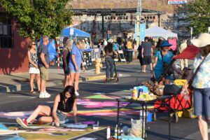 Busy street in the historic district of downtown Grants Pass during AATR 2021. There is a stage with live music, tables with food, and artists drawing on the street with chalk.