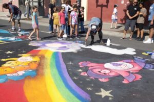 Spectators stand behind an artist that is making chalk art in the street, the art is of Care Bears and a rainbow, from the 80s theme of AATR 2022.