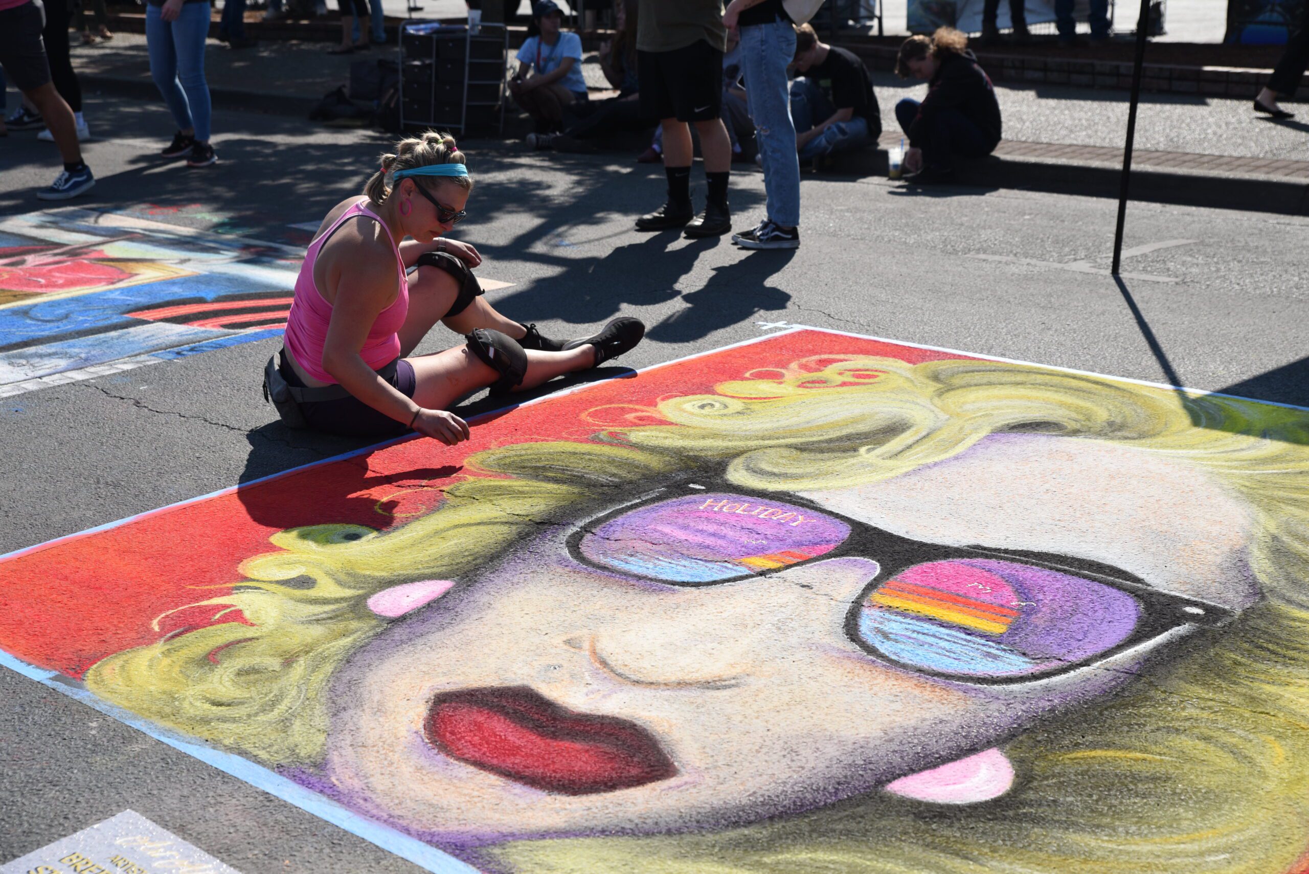 AATR 2022 artist is sitting by a portrait on the street in downtown Grants Pass. The portrait is an old school design of a woman with curly blonde hair, red lipstick, and sunglasses reflecting the setting sun behind the ocean.