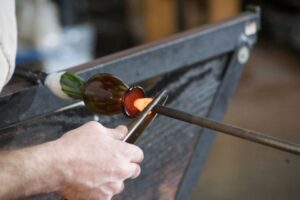 Hand clamping a tool against a metal rod that has a bulb of glowing glass being forged in the workshop at the Glass Forge in Grants Pass.