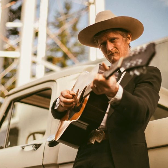 Jeremy James holding a guitar, wearing a cowboy hat and looking at the camera, standing in front of an old school white truck.