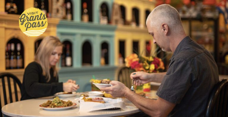 A couple is dining at Cultured Palate, sitting across from each other, in the background there are teal and yellow shelves full of bottles of wine.