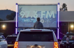 Cars gather in front of a large screen, supported by two metal fixtures with purple neon lights. One person sits on the roof of their SUV to get a clear view of the screen.