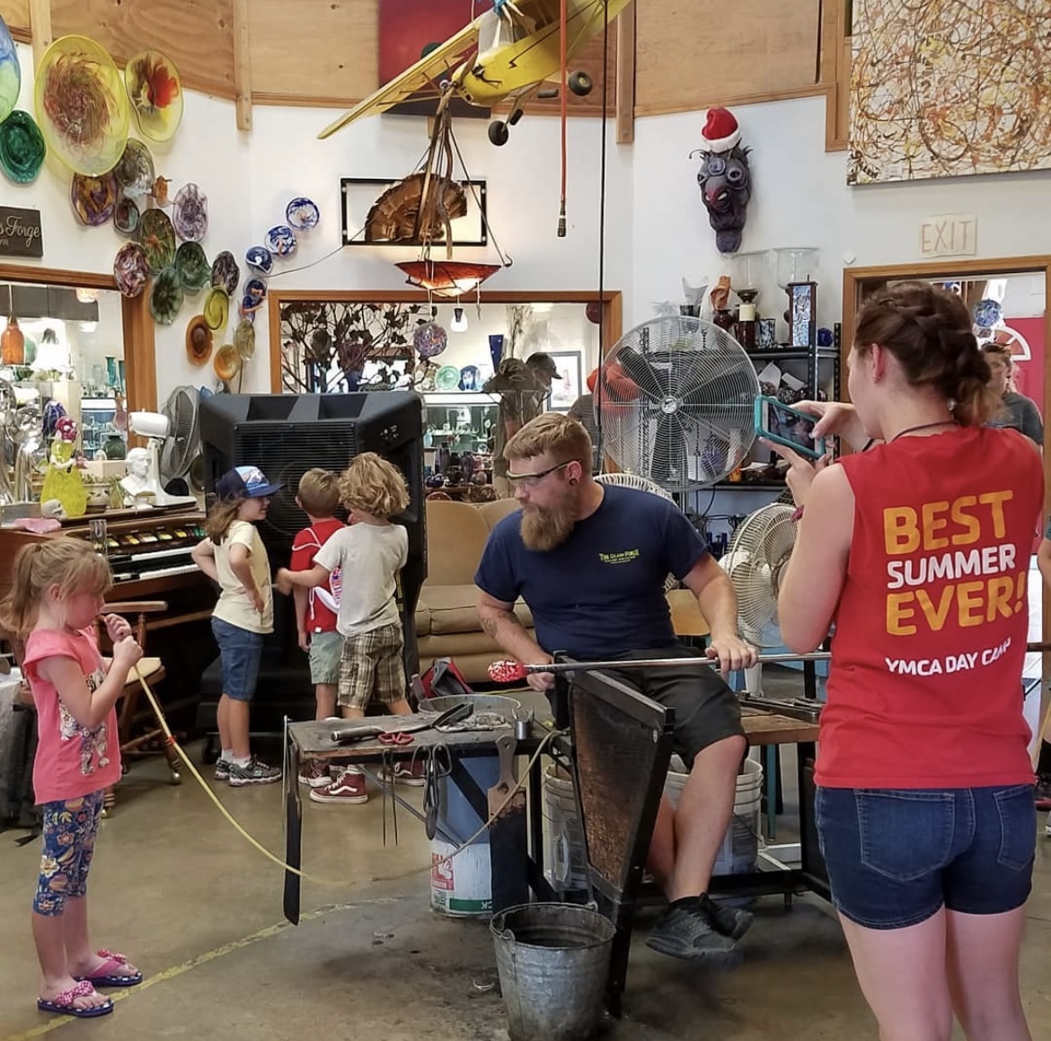 Kids gather around the workshop at the Glass Forge, some admiring the artwork already created, while another participates with one of the craftsman while blowing glass.