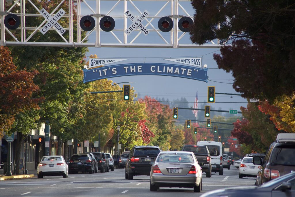 Downtown Grants Pass busy street under the historic "It's the Climate" sign. It is a Fall day with dark red, orange, yellow, and green in the trees.