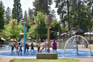 Kids play in an outdoor water park with fountains, sprinklers, and a wooden barrel on top of a pole. A great outdoor activity for the kids in Grants Pass.