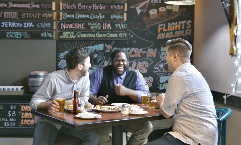 A group of three people laugh together as they sit and enjoy a beer and pizza in Wild River Brewing. A draft list of local craft beers is visible on the chalkboard behind them.