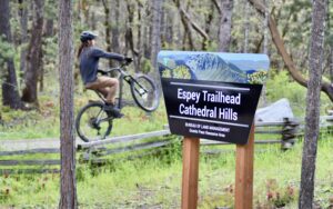 A mountain biker is doing a wheelie on the Espey Trail, as they pass the trailhead sign, located in Cathedral Hills, Grants Pass.